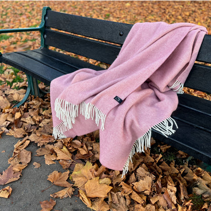 Pink blanket with white fringe draped over a black bench surrounded by autumn leaves.